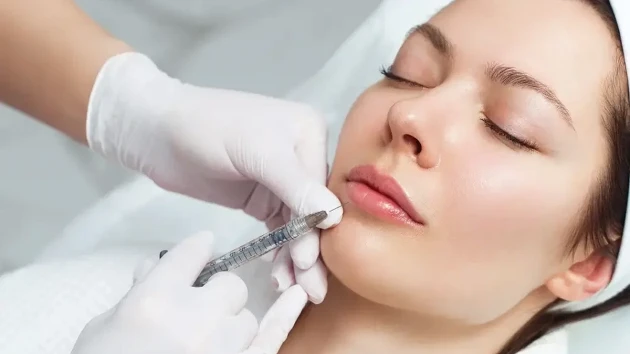 دختری که چشم های خود را بسته و پزشک به چانه و زیر لب او هیالورونیک اسید تزریق می کند